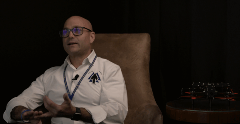 Chris Grollnek, is a man wearing glasses and a white collared shirt sits in a brown chair against a dark backdrop, speaking with expressive hand gestures. A small drone with red propeller blades rests on a round glass table beside him.